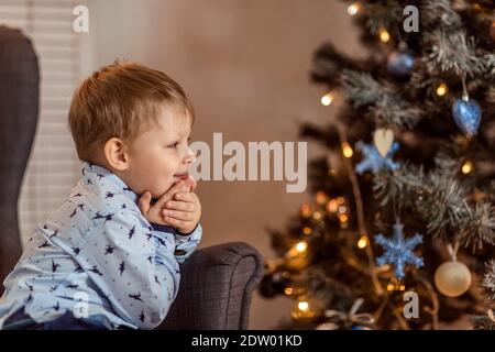 Ein schöner Junge von 4-5 Jahren steht (sitzt) in der Nähe des Sofas und träumt von einem Urlaub und Geschenke. Das Konzept der Winterferien. Selektiver Weichfokus, Stockfoto