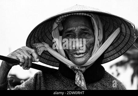 Hoi-An, Vietnam - 9. Dezember. 2001: Porträt einer alten armen Bäuerin mit traditionellem kegelförmigen Reishut, der schwere Lasten mit Stock trägt Stockfoto