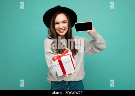 Schuss von ziemlich lächelnd positive junge Brünette Frau isoliert über Blaue Hintergrund Wand trägt stilvolle schwarzen Hut und grauen Pullover Geschenkschachtel halten Stockfoto