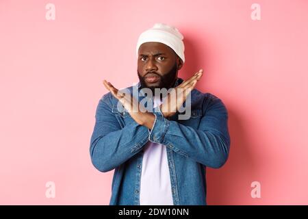 Enttäuscht Schwarzer Mann, starrte auf Kamera unzufrieden und runzelte die Stirn, zeigt Stop Cross Geste, verbieten und nicht einverstanden, stehen über rosa Hintergrund Stockfoto