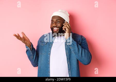 Schöner moderner afroamerikanischer Mann, der auf dem Handy spricht, lächelt und etwas diskutiert, über rosa Hintergrund stehend Stockfoto