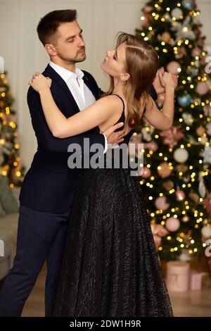 Schöne Paar tanzen im Wohnzimmer. Frau in schwarzem Kleid und Mann in Anzug Umarmung in der Weihnachtszeit. Stockfoto