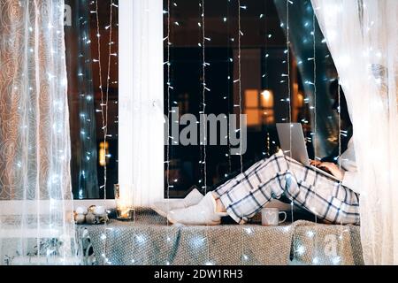 Frau in Pijamas sitzt auf der Fensterbank mit einem Laptop-Computer, der von zu Hause aus arbeitet. Winterstimmung. Weihnachtsbeleuchtung. Stockfoto