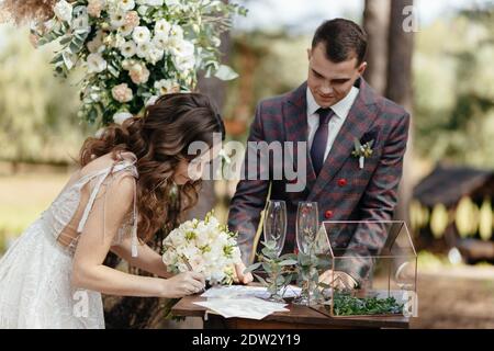 Lächelnde Braut, die Hochzeitspapiere unterschreibt, während der Bräutigam sie anschaut. Außerhalb der Hochzeitszeremonie. Glückliches Jungvermählte bei der Hochzeitszeremonie Stockfoto