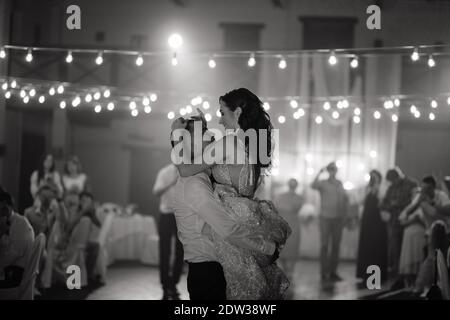 Bräutigam hält die lächelnde Braut in den Händen. Fröhliches Brautpaar, das auf der Hochzeitsfeier tanzt. Frisch verheiratetes Paar tanzt zuerst. Schwarz und Weiß Stockfoto