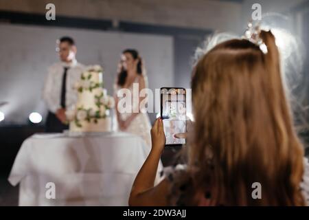 Kleines Mädchen, das ein Foto von Brautpaar und Kuchen auf dem Smartphone. Ort für Ihren Text. Hintergrund der Hochzeit Stockfoto