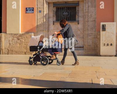 Kaukasische Frau, die mit dem Kinderwagen und ihrer Tochter auf dem Kutschenzubehör durch die Straßen der Stadt geht. Beide tragen Masken. Stockfoto