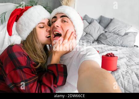 Online-Weihnachtsfeier. Frau beißende Mann nimmt Selfie trägt santa Hüte und roten Pyjamas vor dem Hintergrund der Neujahrsgeschenke Stockfoto