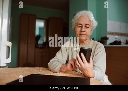 Porträt einer älteren Großmutter, die Nachrichten an ihre Familie am Telefon tippt, während sie an einem Tisch im Haus sitzt. Kommunikation während der Isolation Stockfoto