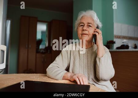 Porträt einer älteren Großmutter, die mit ihrer Familie telefoniert, während sie an einem Tisch im Haus sitzt. Kommunikation während der Isolierung von COVID-19 Stockfoto