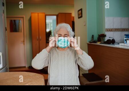 Porträt einer älteren Frau, die eine Gesichtsmaske aufsetzt, während sie an einem Tisch im Haus sitzt. Prävention der Gesundheit älterer Menschen während der Isolation Stockfoto