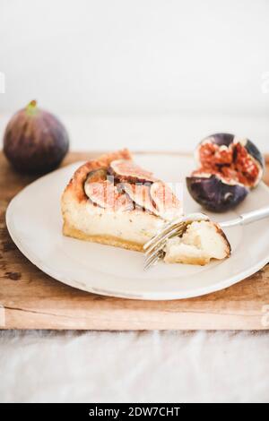 Scheibe hausgemachter Feigenkäsekuchen mit frischen Feigen auf weißem Teller über rustikalem Holzbrett, weiße Wand im Hintergrund, Nahaufnahme. Gesundes Dessert, handwerklich Stockfoto