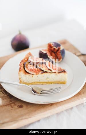 Scheibe hausgemachter glutenfreier Feigenkäsekuchen mit frischen Feigen der Saison auf weißem Teller über rustikalem Holzbrett, weiße Wand im Hintergrund. Gesundheit Stockfoto