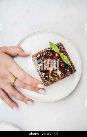 Flaches Lay der Frau, die Kakao-Tartalette mit frischen saisonalen Beeren von Hand aus weißem Teller nimmt, Draufsicht. Modernes Dessertkonzept Stockfoto