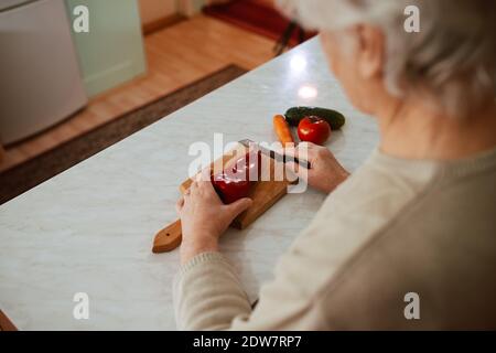 Blick von der Rückseite einer Großmutter, die das Mittagessen kocht und Gemüse auf dem Küchentisch zerkleinert. Eine grauhaarige Rentnerin beim Kochen des Mittagessens Stockfoto