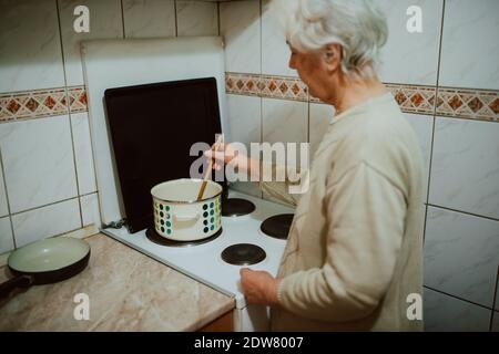Blick von der Rückseite einer älteren Frau beim Kochen Mittagessen in der Küche. Eine pensionierte grauhaarige Großmutter neben einem Küchenherd Stockfoto