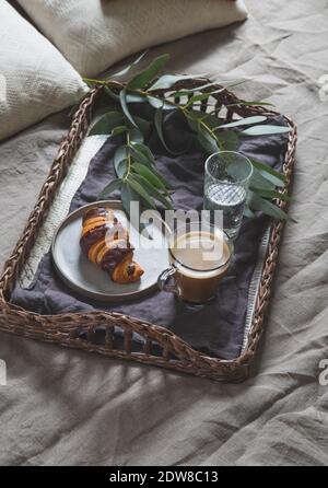 Tablett mit Korbblech und Kaffee und Croissant auf einem Leinenbett. Frühstück im Bett. Stockfoto