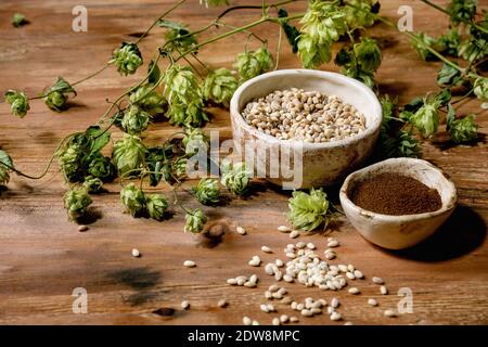 Alles zum Brühen. Frische grüne Hopfenzapfen, Weizenkorn und rotfermentiertes Malz in Keramikschalen auf Holzhintergrund. Stockfoto