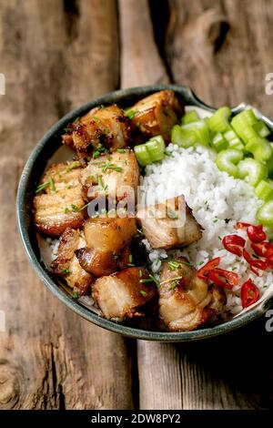 Gegrillter frittierter Schweinebauch in einer Schüssel mit Reis, Sellerie, Chili und Frühlingszwiebeln auf altem Holzhintergrund. Stockfoto