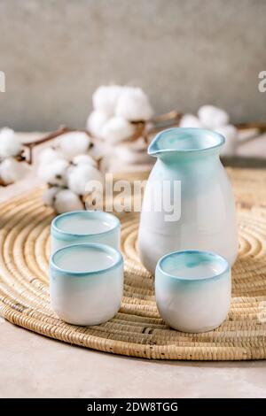 Sake Keramik Set für traditionelle japanische Alkohol trinken Reiswein Sake, Krug und drei Tassen, stehend auf Strohserviette mit Baumwollblumen über beig Stockfoto