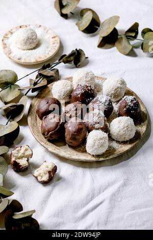 RAW vegan hausgemachte Kokosschokolade Süßigkeiten Kugeln mit Kokosflocken in Keramik-Platte über weißem Textil-Hintergrund mit Eukalyptus-Zweigen. Stockfoto
