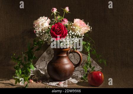 Stillleben mit einem Strauß Rosen und Gypsophila in einem Krug und einem Apfel auf einem Hintergrund von Sackleinen. Selektiver Fokus Stockfoto