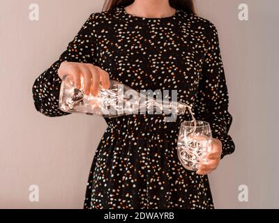 Gesichtslose Schuss der Frau in festlichem Kleid hält Glas Becher und Gießen glühende Girlande aus Weinflasche. Winterferien, Weihnachtsfestliches Konzept. Stockfoto