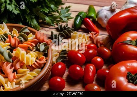 Terracotta-Schüssel mit tricolor Fusilli Pasta und Kirsche und runde Tomaten, Chilischoten, Knoblauch und Petersilie. Konzept der Lebensmittelrohstoffe. 45 Grad Winkel Stockfoto