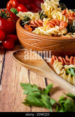 Terracotta-Schüssel mit dreifarbigen Fusilli-Nudeln und Kirsche und runde Tomaten, Chili, Knoblauch und Petersilie. Konzept der Lebensmittelrohstoffe. 45-Grad-Winkel Stockfoto