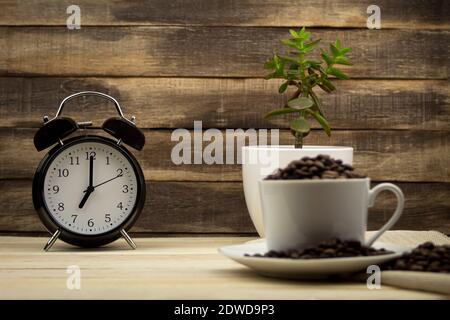 Der Wecker zeigt sieben Uhr an. Weiße Tasse mit Kaffeesorten auf dem Hintergrund der Uhr. Holzhintergrund Stockfoto