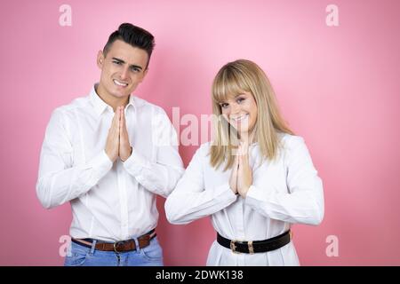 Junges Paar von Freundin und Freund über isoliert rosa Hintergrund Betteln und Beten mit Händen zusammen mit Hoffnungsausdruck auf Gesicht sehr emoti Stockfoto