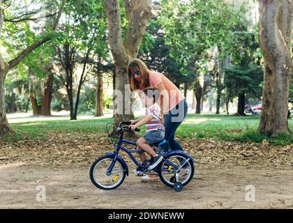 Mutter lehrt ihrem kleinen Sohn, ein Fahrrad zu fahren. Kleiner Junge auf einem blauen Fahrrad in einer schmutzigen Straße in einem Waldpark und half von seiner Mutter. Fahrrad mit Th Stockfoto