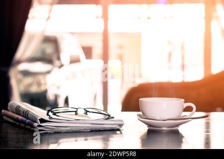 Nahaufnahme Tasse heißen Cappuccino Kaffee mit Zeitung und Brille auf dem Holztisch mit Blur Coffee Shop Hintergrund-warmen Ton. Stockfoto