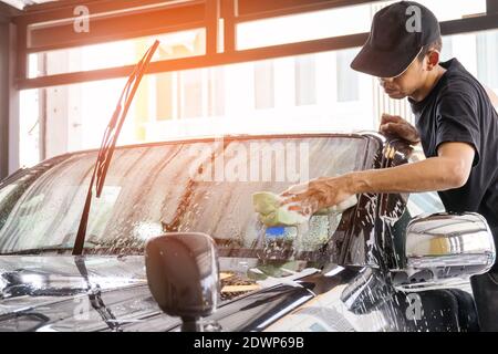 Autowascharbeiter mit T-Shirt und schwarzer Kappe reinigen das Auto in der Autowaschanlage mit einem Schwamm, Konzept für die Autopflege. Stockfoto