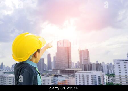 Ein Junge, der als Ingenieur gekleidet ist und einen Schutzhut trägt Zeigt seine Hand auf das Stadtbild mit Sonnenuntergang Hintergrund-Konzepte für Die Zukunft wächst bis zu b Stockfoto