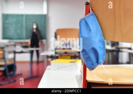 Leeres Klassenzimmer, die Stühle stehen auf dem Schreibtisch - eine Gesichtsmaske, Maske hängt auf einem Stuhl - ein Lehrer steht vor der Tafel im Hintergrund. Schulen und Kindertagesstätten werden am 21. Dezember 2020 aufgrund der auferlegten Sperrung für eine zunächst unbestimmte Zeit geschlossen, Klassenzimmer einer Grund- und Mittelschule in Germering bei München in Bayern. Weltweite Nutzung Stockfoto