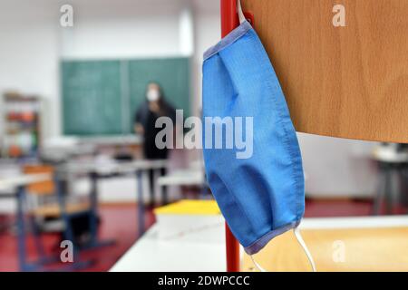 Leeres Klassenzimmer, die Stühle stehen auf dem Schreibtisch - eine Gesichtsmaske, Maske hängt auf einem Stuhl - ein Lehrer steht vor der Tafel im Hintergrund. Schulen und Kindertagesstätten werden am 21. Dezember 2020 aufgrund der auferlegten Sperrung für eine zunächst unbestimmte Zeit geschlossen, Klassenzimmer einer Grund- und Mittelschule in Germering bei München in Bayern. Weltweite Nutzung Stockfoto