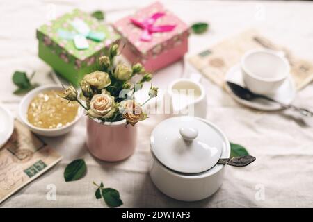 Essen Stillleben im französischen Stil. Weiße Kaffeetassen, Milchkännchen mit Sahne, Rosen und zwei Geschenken auf einer Leinentischdecke. Verfeinerte Tonung. Stockfoto