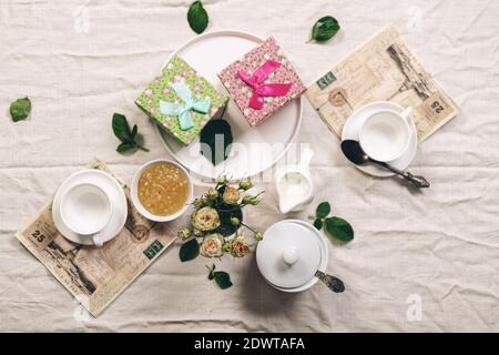 Essen Stillleben im französischen Stil. Weiße Kaffeetassen, Milchkännchen mit Sahne, Rosen und zwei Geschenken auf einer Leinentischdecke. Verfeinerte Tonung. Draufsicht Stockfoto