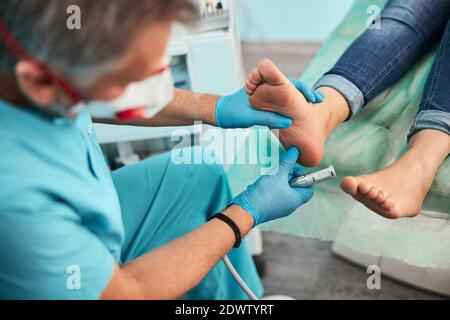 Schöner Mann in blauen Handschuhen, der den linken Fuß in der Hand hält Während harte Haut Peeling im medizinischen Zentrum Stockfoto