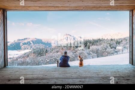 Frau mit Thermoskanne sitzt mit ihrem Beagle Hund in Der große Holzhangar mit einem riesigen Panoramafenster und Blick auf die verschneite Berglandschaft Stockfoto