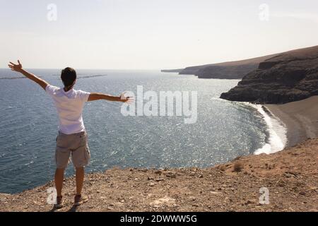 Junger Mann mit offenen Armen auf einer Klippe und Blick auf das Meer auf Lanzarote. Männliche Touristen mit Blick auf vulkanische Küste an sonnigen Tag auf den Kanarischen Inseln Stockfoto