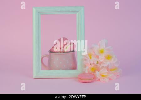 Eine schöne Blumenaufnahme mit Makronen in einem Becher In der Nähe eines Fotorahmens auf einem rosa Hintergrund - noch Leben Stockfoto