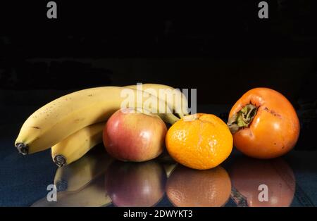 Die Banane, Apfel, Mandarine und Kakimmon auf dem Tisch mit Reflexion. Reife Früchte auf schwarzem Hintergrund Stockfoto