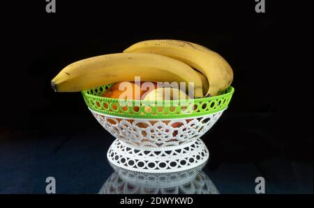 Die Bananen mit Mandarine und Apfel liegen in Vase auf Glastisch. Reife Früchte mit Reflexion auf schwarzem Hintergrund Stockfoto