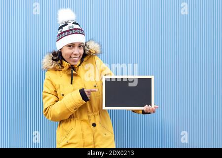 Portrait eines hispanischen Mädchens, das Winterkleidung trägt und auf eine leere Tafel zeigt. Sie steht neben einer blauen Wand. Leerzeichen für Text. Stockfoto