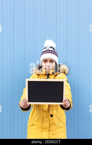 Vertikales Porträt einer ernsthaften lateinischen Frau in Winterkleidung mit einer leeren Kreidetafel. Sie steht neben einer blauen Wand. Leerzeichen für Text. Stockfoto