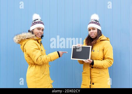 Porträt von zwei lateinamerikanischen Frauen in der gleichen Reihe von Winterkleidung isoliert auf einem blauen Hintergrund gekleidet. Man hält eine leere Tafel, während Stockfoto