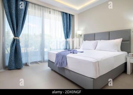 Modernes Design-Interieur des eleganten Schlafzimmers mit weichem grauen Textil-Doppelbett, weißer Bettwäsche und blauen Vorhängen im klassischen Stil Hotel-Apartment Stockfoto