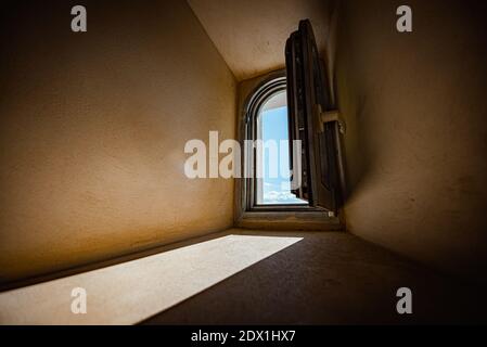 Die Sonne scheint durch ein kleines Fenster im Zimmer mit Steinwänden. Blauer Himmel mit Wolken im Fenster Stockfoto